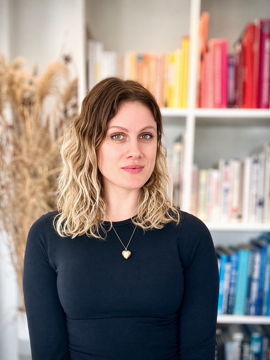 Portrait of Georgie Webb, Tech VA, in front of a colour-organised bookshelf.